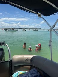 View from a pontoon boat over shallow turquoise coastal water at a sandbar, with several anchored motorboats in the distance and four people wading and chatting under a bright blue sky with scattered clouds.