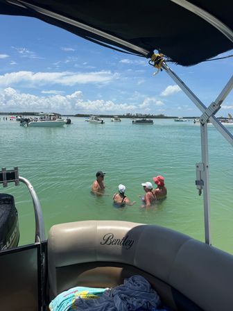 View from a pontoon boat over shallow turquoise coastal water at a sandbar, with several anchored motorboats in the distance and four people wading and chatting under a bright blue sky with scattered clouds.