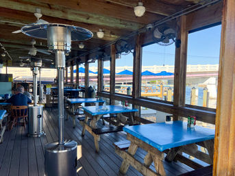 Waterfront dockside outdoor dining deck with blue-topped picnic tables, patio heaters and ceiling fans, blue umbrellas and bridge over the marina