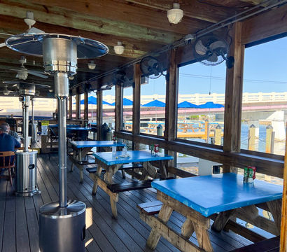 Waterfront dockside outdoor dining deck with blue-topped picnic tables, patio heaters and ceiling fans, blue umbrellas and bridge over the marina