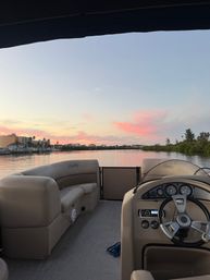 View from a pontoon boat helm and cushioned deck looking out over a calm coastal inlet and marina at sunset, pink clouds, waterfront buildings and palm trees reflecting on the water.