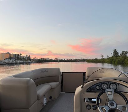 View from a pontoon boat helm and cushioned deck looking out over a calm coastal inlet and marina at sunset, pink clouds, waterfront buildings and palm trees reflecting on the water.