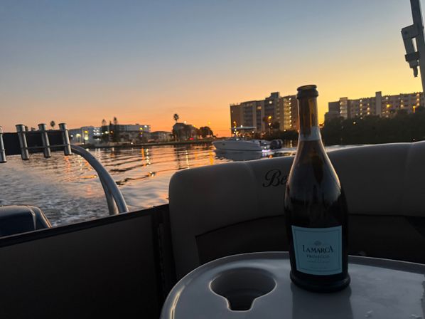 Bottle of prosecco on a pontoon boat table at sunset, calm bay waters reflecting city lights with palm trees and waterfront condos in the background.