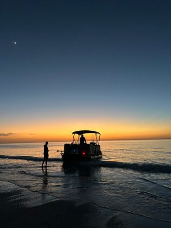 Silhouette of a pontoon boat and a person wading at the shoreline during a vibrant orange-blue coastal sunset with a crescent moon and calm sea reflections.