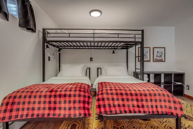 Cozy basement guest bedroom with black metal bunk frame, two twin beds below topped with white linens and red-and-black buffalo plaid blankets, wall-mounted reading lamps, cubby storage and framed animal prints.