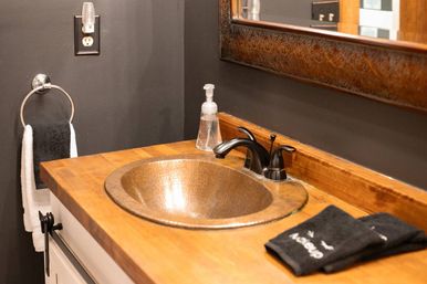 Rustic bathroom vanity with round hammered copper sink set into a wood countertop, black double-handle faucet, clear soap dispenser, folded black hand towels, dark gray wall and ornate framed mirror