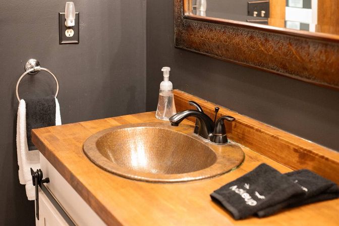 Rustic bathroom vanity with round hammered copper sink set into a wood countertop, black double-handle faucet, clear soap dispenser, folded black hand towels, dark gray wall and ornate framed mirror