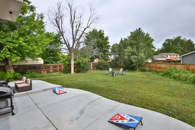 Suburban backyard with concrete patio featuring outdoor sofa and cornhole boards, large grassy yard with Adirondack chairs around a firepit, mature trees and wooden privacy fence under an overcast sky.