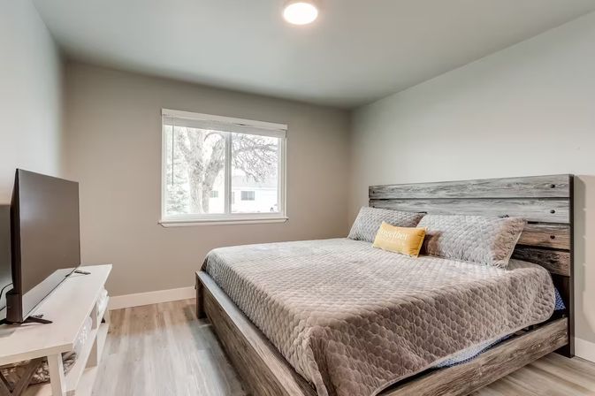 Bright modern bedroom with rustic gray wood platform bed and quilted gray bedding, yellow accent pillow, flat-screen TV on a white stand, large window showing a tree and nearby houses, and light wood flooring.