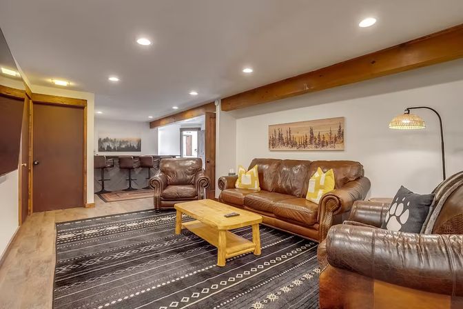Cozy rustic basement living room and rec room with brown leather sofa and armchairs, wooden coffee table on a patterned black area rug, exposed wood beam and recessed lighting, pine-tree wall art, floor lamp, and bar-style seating in the background.