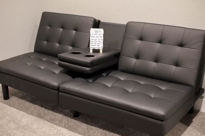 Black tufted faux-leather two-seat convertible sofa bed with a fold-down center armrest and built-in cup holders on a carpeted floor against a beige wall, small handwritten note on the armrest.