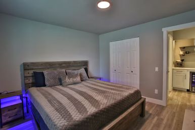 Cozy modern bedroom with king-size bed and rustic wooden headboard, gray quilted bedding, blue LED under-bed lighting, double-door closet and a kitchenette visible through the doorway.