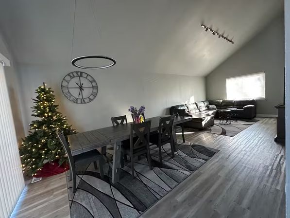 Modern open-plan living and dining room with gray floors and walls, wooden dining table and chairs, lit Christmas tree, large wall clock, leather sectional sofa under a vaulted ceiling and bright window.
