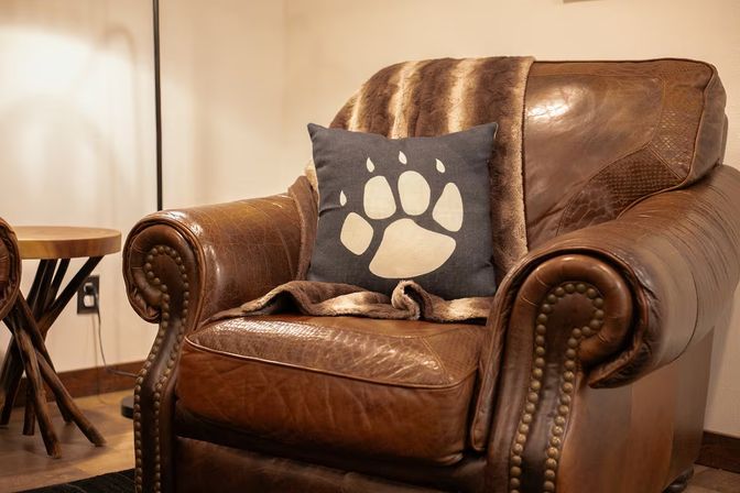 Cozy brown leather armchair in a rustic living room, styled with a navy pillow featuring a cream paw-print, a faux-fur throw draped over the back, wooden side table and warm ambient lighting.