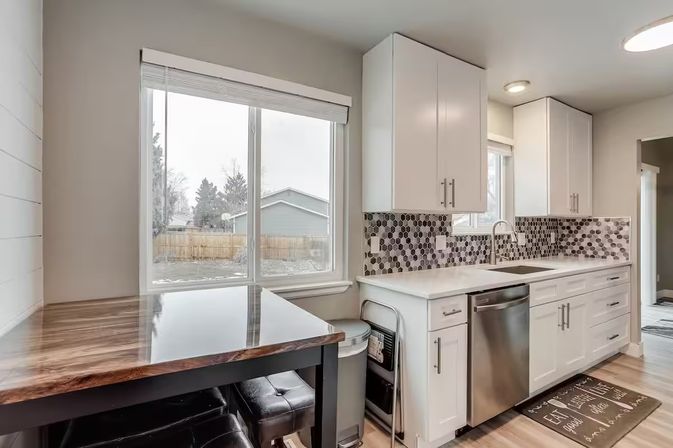 Bright modern suburban kitchen with white cabinets, hexagon tile backsplash, stainless dishwasher and sink, wood-topped breakfast bar with leather stools under a large window overlooking a fenced backyard.
