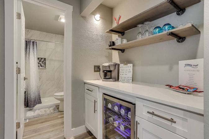 Cozy modern guest kitchenette and wet bar with glass-front wine fridge, coffee maker, open wood shelves with glassware and blue décor, countertop welcome note and snacks, and an adjacent bathroom with tiled shower and ombré curtain.