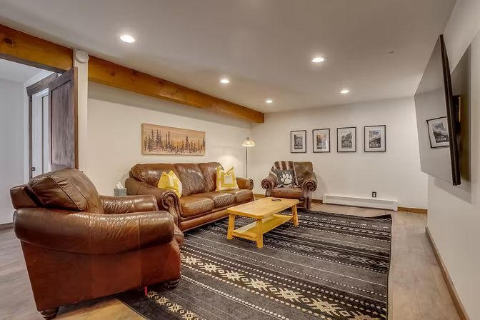 Cozy rustic living room with brown leather sofa and armchairs, patterned black rug, wooden coffee table, exposed ceiling beam, recessed lights, and wall-mounted TV.