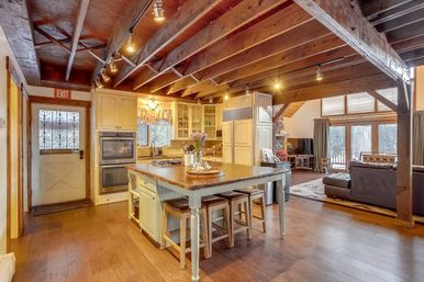 Warm rustic open-concept cabin interior with exposed wood beams, central kitchen island and stools, double oven and farmhouse door, and a cozy living area with large windows overlooking snowy trees.