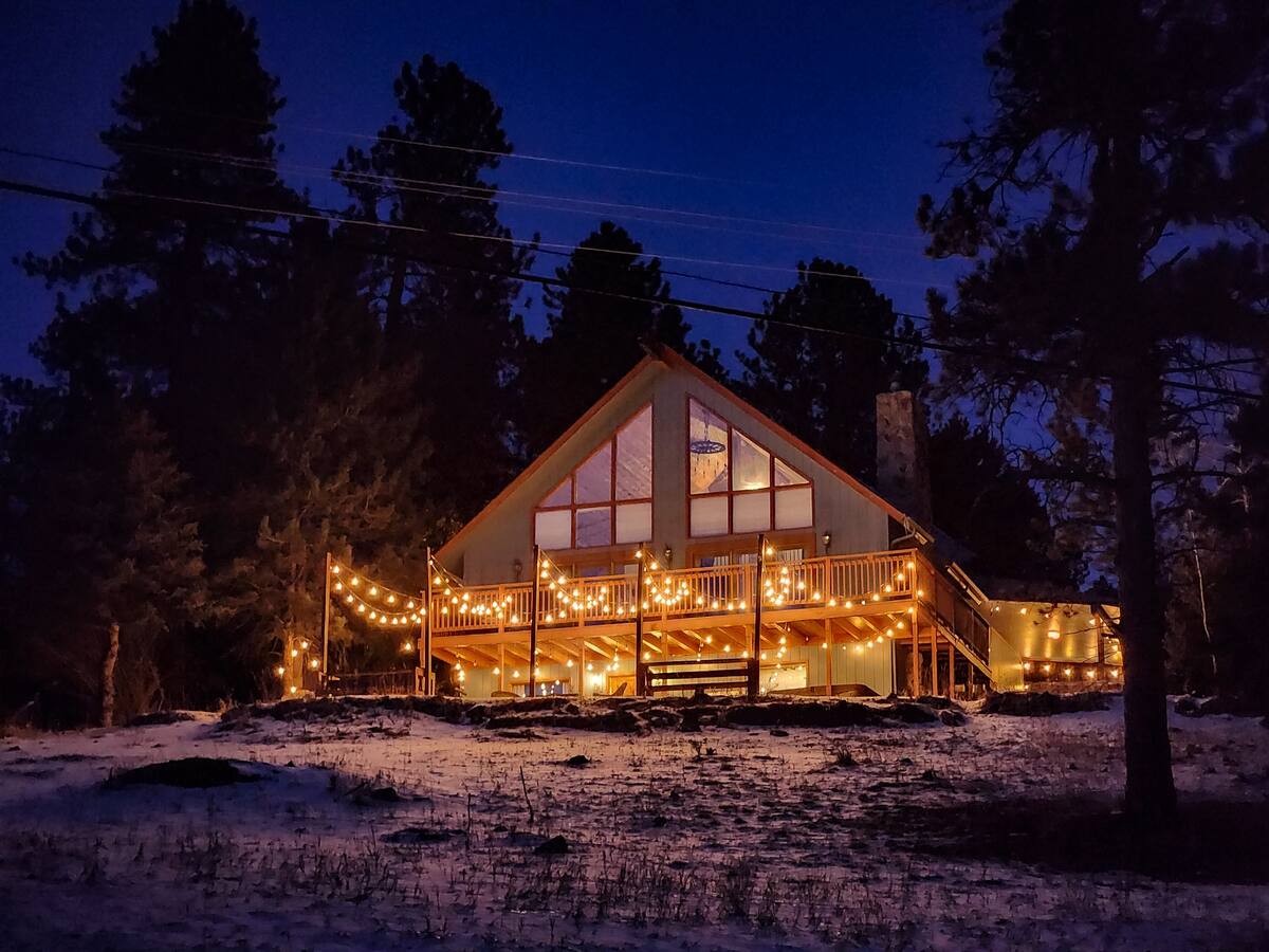Cozy A-frame mountain cabin in a snowy pine forest at dusk, warm string lights wrapped around a wooden deck and glowing through large windows against a deep blue sky.