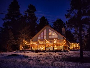 Cozy A-frame mountain cabin in a snowy pine forest at dusk, warm string lights wrapped around a wooden deck and glowing through large windows against a deep blue sky.