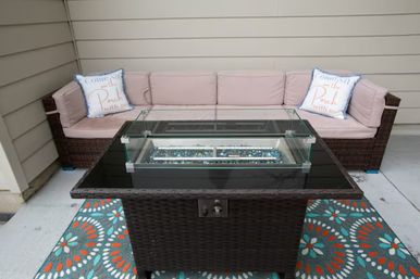 Cozy outdoor patio lounge with dark wicker sectional and blush cushions, decorative pillows, central glass-top fire pit table with blue fire glass, and a colorful floral outdoor rug against beige siding.
