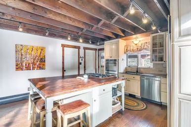 Rustic farmhouse kitchen with exposed dark wood ceiling beams, a polished wooden island with leather stools, white cabinets, stainless steel appliances, track lighting, and birch tree wall art.