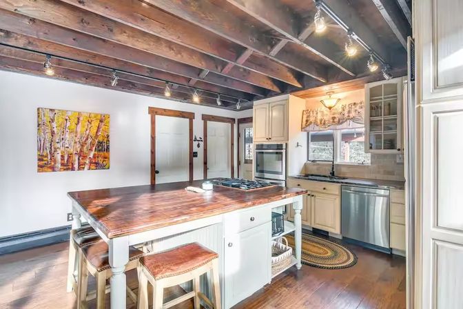 Rustic farmhouse kitchen with exposed dark wood ceiling beams, a polished wooden island with leather stools, white cabinets, stainless steel appliances, track lighting, and birch tree wall art.