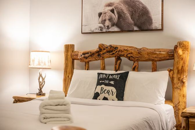 Cozy rustic mountain cabin bedroom with handcrafted log bedframe, bear-themed pillow reading Don't wake the bear, framed bear photo above the headboard, stacked white towels and antler-style bedside lamps.