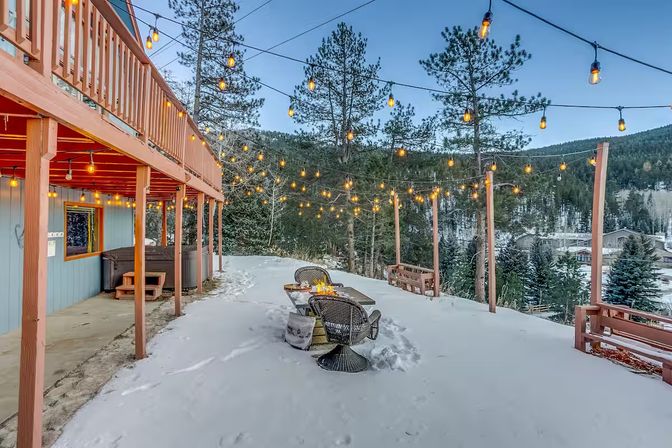 Cozy snow-covered mountain deck at dusk with hanging string lights, wicker chairs around a fire-pit table, hot tub beneath a raised wooden balcony and pine-forested valley views.