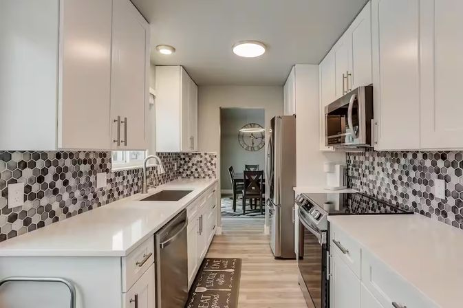 Bright modern galley kitchen with white shaker cabinets, quartz countertops, gray hexagonal tile backsplash, stainless steel appliances, undermount sink and wood-look plank flooring opening to a dining area with a wall clock.