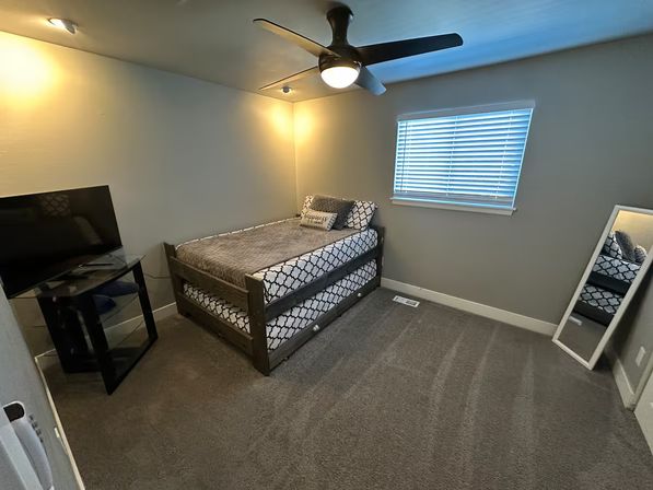 Small modern gray guest bedroom with a trundle daybed in patterned bedding, ceiling fan with light, TV on a glass stand, full-length leaning mirror, carpeted floor and window blinds