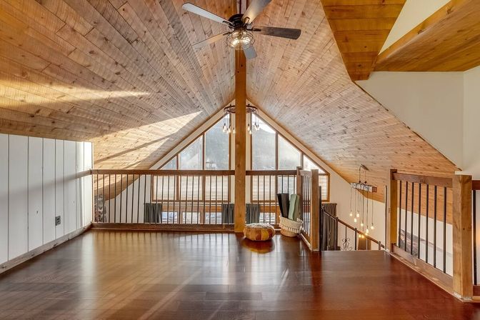 Rustic cabin loft with vaulted wood-paneled ceiling, dark hardwood floors, ceiling fan, wooden railing and large triangular windows flooding the bright living area with natural light.