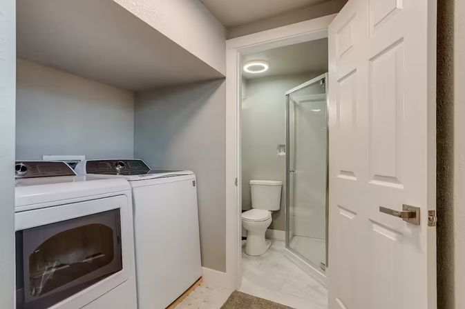 Compact modern laundry room with side-by-side white washer and dryer opening to an adjoining bathroom with a glass shower, toilet, and neutral gray walls.