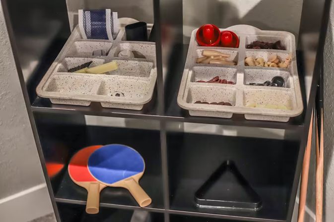 Game-room cubby shelf with speckled compartment trays holding small parts and two red table tennis balls, two red-and-blue ping-pong paddles, a black pool triangle rack and wooden cue sticks.