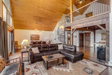 Rustic mountain-cabin loft living room with vaulted pine ceiling, large windows, dark leather sectional on a Southwestern patterned rug, wooden coffee table, spiral staircase to a mezzanine, and dining area in back.