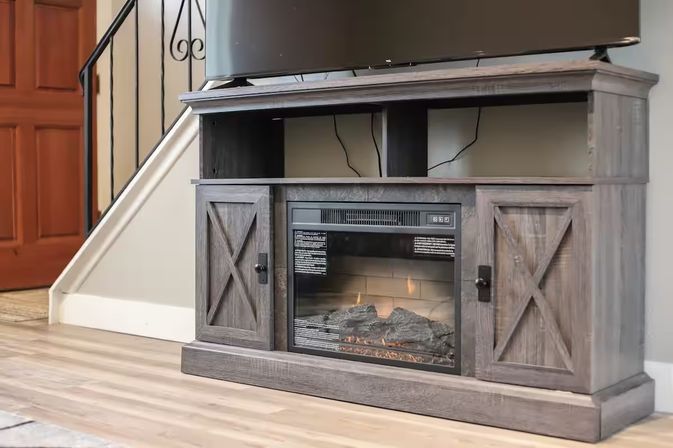 Cozy living room with a rustic gray farmhouse media console featuring a built-in electric fireplace and flat-screen TV on hardwood floors beside a staircase and entry door.