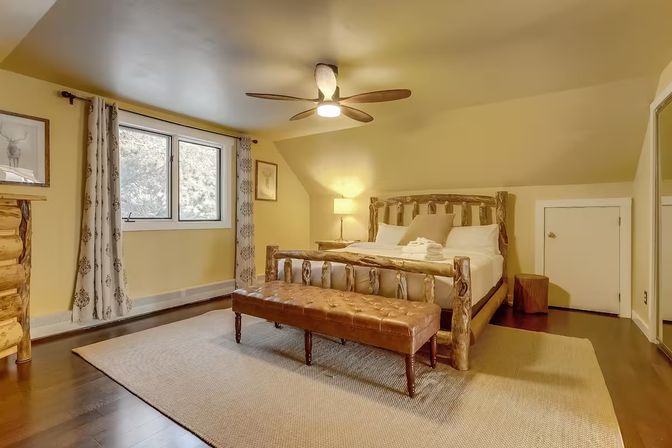 Cozy rustic bedroom with a log-frame bed, tufted leather bench at the foot, ceiling fan, patterned curtains, dark hardwood floors and warm yellow walls.
