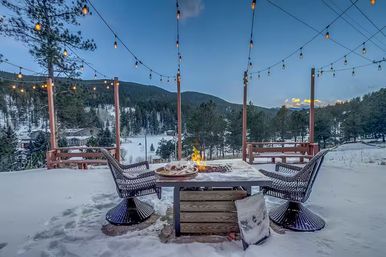Snow-covered mountain deck at dusk with hanging string lights, two wicker chairs around a glowing fire-pit table set with plates, overlooking pine trees and snowy hills.
