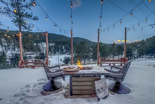 Snow-covered mountain deck at dusk with hanging string lights, two wicker chairs around a glowing fire-pit table set with plates, overlooking pine trees and snowy hills.