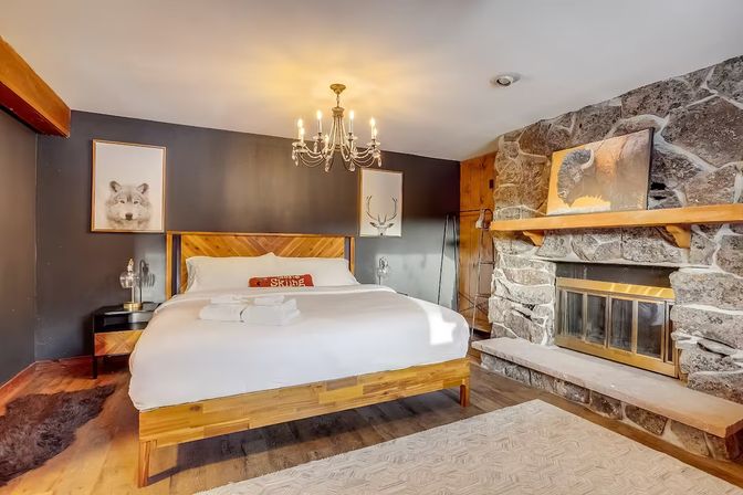 Cozy rustic mountain cabin bedroom with a king bed and wooden headboard, stone fireplace with mantel, chandelier lighting, wildlife wall art, hardwood floors and warm, inviting decor.