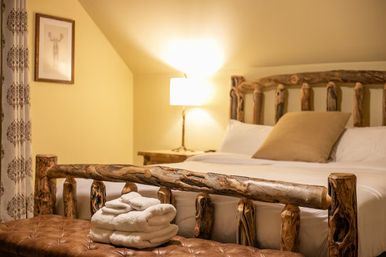 Cozy rustic mountain-cabin bedroom with handcrafted log bed, white linens and tan pillow, tufted leather bench topped with folded white towels, warm bedside lamp and patterned curtains.