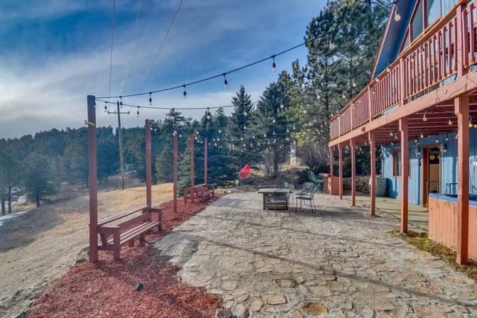 Stone patio at a blue A-frame mountain cabin with wooden deck and benches, overhead string lights, metal table and chairs, and pine-forested hillside views