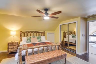 Cozy rustic mountain-cabin bedroom with log-frame bed, patterned throw and green pillows, tufted leather bench, mirrored closet doors, ceiling fan and balcony access.