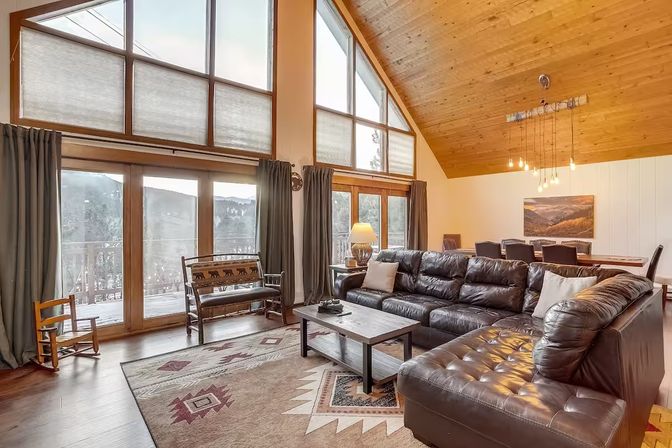 Cozy mountain-cabin living room with vaulted wood ceiling, floor-to-ceiling windows and balcony views, large dark leather sectional, rustic patterned rug, wooden coffee table and adjacent dining area.