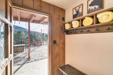 Rustic mountain cabin entryway with wooden bench, coat hooks and rolled yellow towels, opening onto stone patio with string lights and pine-tree mountain view.