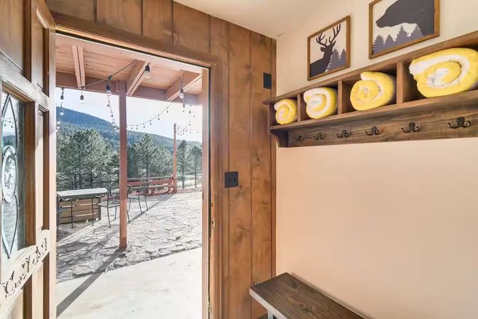 Rustic mountain cabin entryway with wooden bench, coat hooks and rolled yellow towels, opening onto stone patio with string lights and pine-tree mountain view.