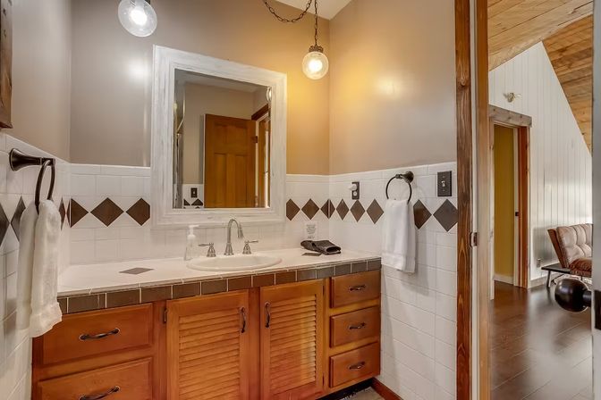 Cozy rustic bathroom with wooden louvered vanity, white tile backsplash with brown diamond accents, framed mirror, globe pendant lights, chrome faucet and towel rings, and an open doorway to a wood-paneled room.