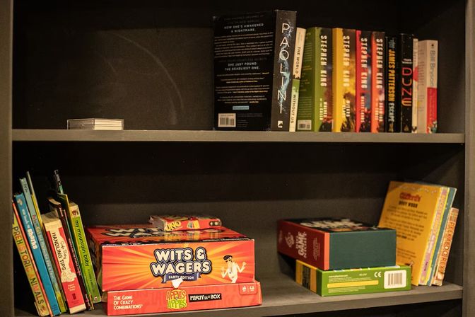 Home bookshelf in a living room with colorful board games and books, Wits & Wagers party box centered, kids' picture books on the left and paperback novels on the top shelf.