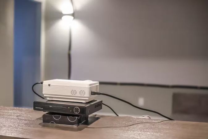 White tabletop projector stacked on a black media player and small box on a wooden table, cables plugged in, with a floor lamp and blurred projection screen in the background — cozy home theater setup.