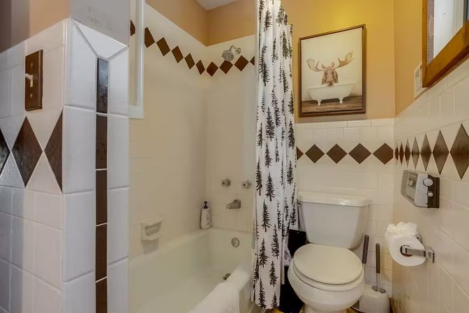 Cozy cabin-style bathroom with a bathtub-shower combo, pine tree patterned shower curtain, white tiles with brown diamond trim, white toilet and framed moose-in-a-tub artwork.
